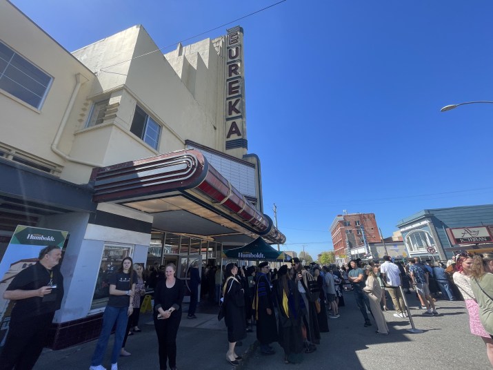 Graduates wearing black gowns and caps, friends, and family crowd around an old fashioned theater with beige paint and neon letters spelling "Eureka" on the outside.