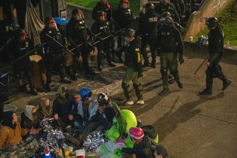 A group of students and community members, most wearing masks and some wearing helmets, sit in a circle on the ground in the bottom-left corner. They are surrounded by officers in riot gear like black helmets, vests, and holding batons.